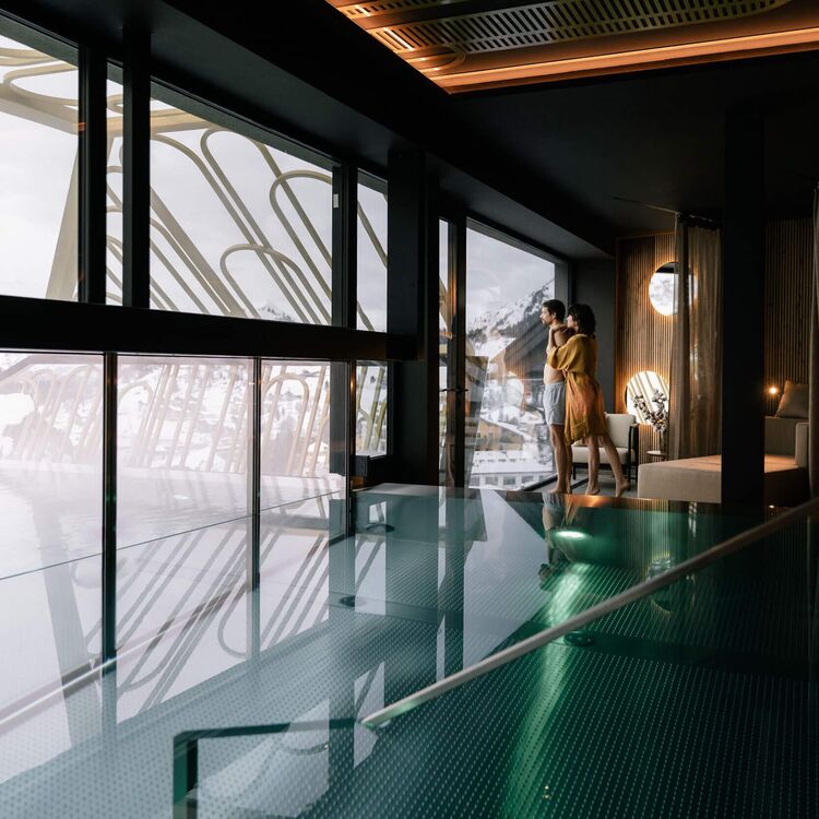 Ein Paar genießt das Gästeerlebnis in einem modernen Hotelzimmer mit Blick auf alpine Architektur und gläsernem Pool.