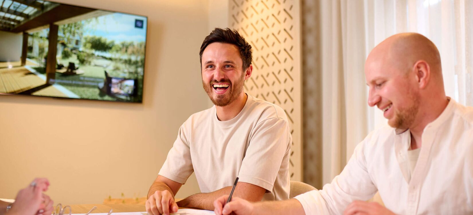 Zwei Männer lächeln, während sie an kreativen Hotelkonzepten und Planungen in einem modernen Raum arbeiten.