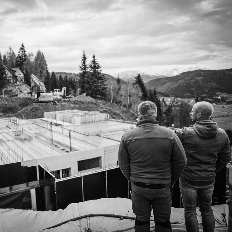 Zwei Männer beobachten die Fortschritte beim Hotelneubau in malerischer alpiner Landschaft.
