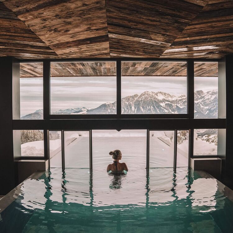 Eine Person entspannt im Pool eines modernen Hotels mit alpiner Architektur und Blick auf die Berge.